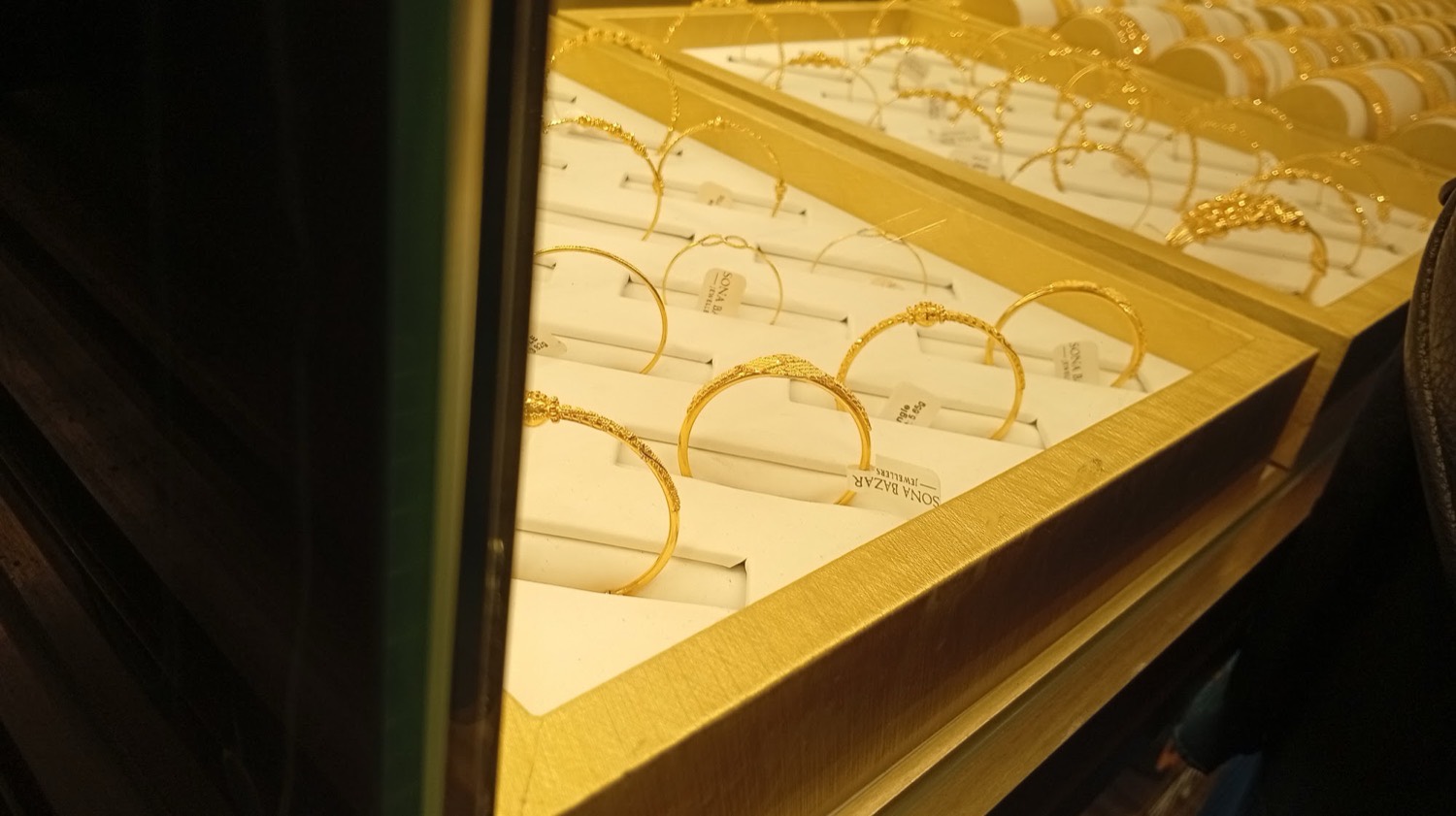 Patterned gold bangles, each tagged, laid out in a lit counter tray at Sona Bazar Jewellers