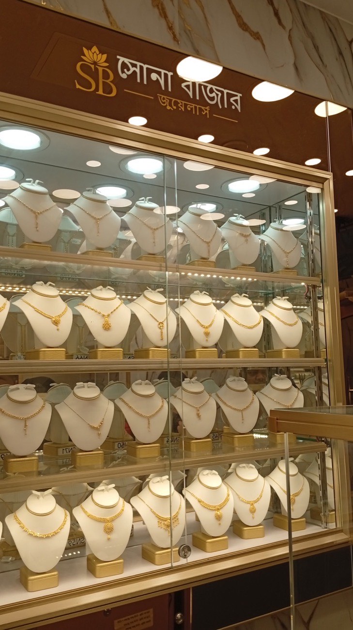 A tall glass cabinet full of gold necklace sets on white busts at Sona Bazar Jewellers, beneath the Bengali shop sign